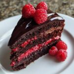 A decadent slice of chocolate raspberry cake featuring dark layers, bright raspberry filling, and topped with chocolate ganache and fresh raspberries.