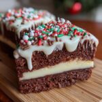 Close-up of a slice of chocolate christmas tree cheesecake with white frosting and festive sprinkles.
