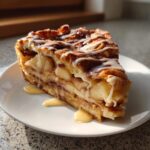 A close-up slice of cinnamon roll apple pie, layered with apples and topped with white icing.