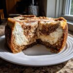 A rich, creamy cinnamon roll cheesecake with a piece removed, showing the swirl of cream cheese and cinnamon filling.