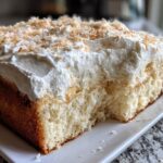 Close-up of a moist slice of coconut poke cake topped with thick white frosting and toasted coconut flakes.