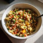 A white bowl filled with vibrant cauliflower rice mixed with scrambled egg, diced carrots, peas, and green onions.
