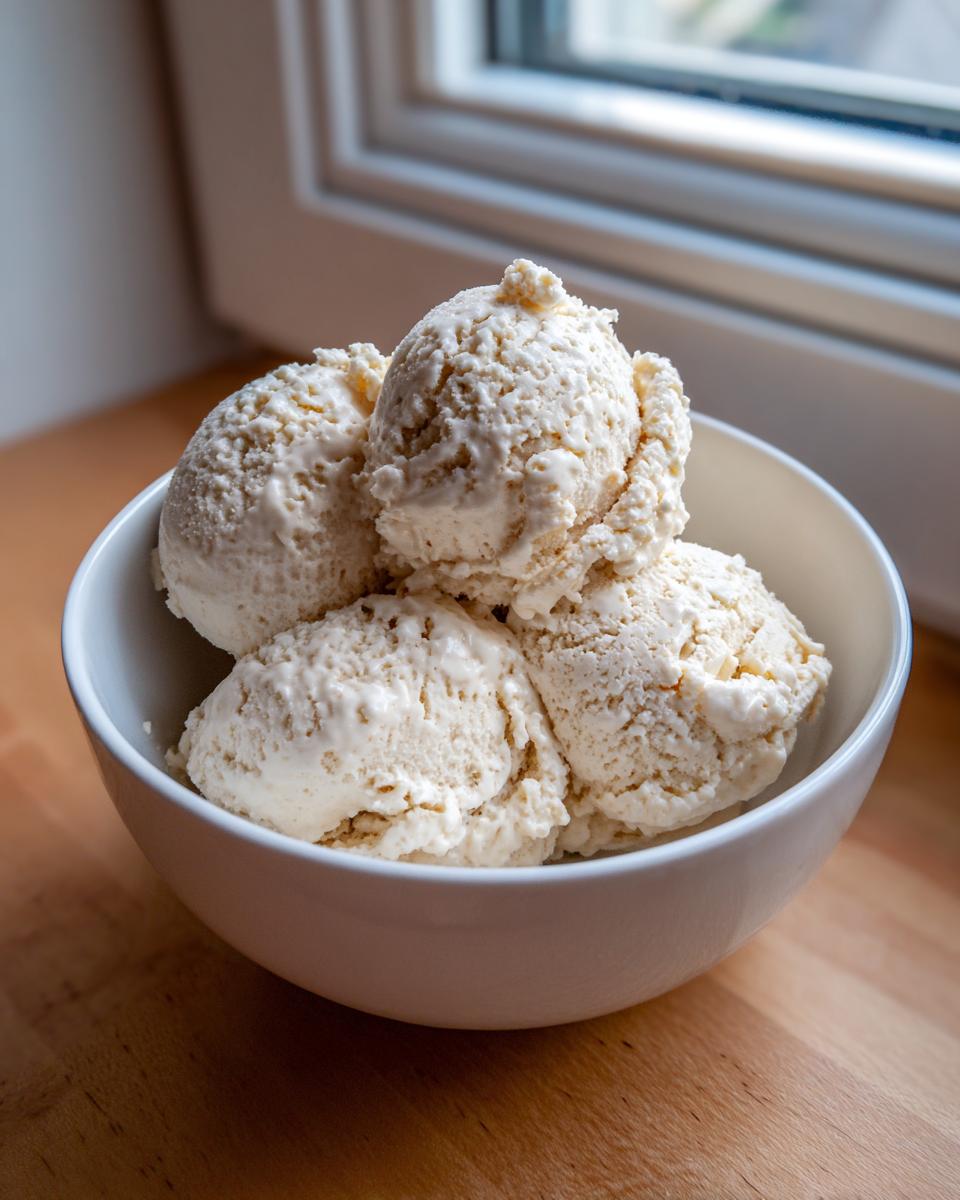 Four scoops of creamy, vanilla-colored cottage cheese ice cream served in a white bowl on a wooden surface.