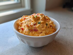 A white bowl filled with creamy, orange cottage cheese queso dip mixed with visible red peppers and green herbs.