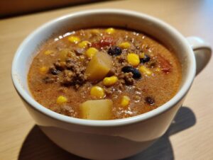 Close-up of a white mug filled with rich, reddish-brown cowboy soup containing ground beef, corn, potatoes, and black beans.