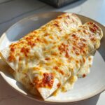 Two creamy chicken enchiladas covered in bubbly, browned melted white cheese, served on a light plate.
