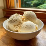 Three scoops of creamy, pale yellow cottage cheese ice cream served in a white bowl on a wooden surface.