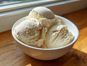 Three scoops of creamy, pale yellow cottage cheese ice cream served in a white bowl near a window.
