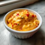 Close-up of creamy, orange cottage cheese queso mixed with diced red peppers in a small white bowl.