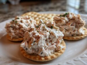 Three servings of creamy ham salad recipe piled high on round crackers, garnished with herbs, on a white plate.