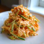 A mound of creamy, shredded kani salad mixed with thin strips of green cucumber served on a white plate.