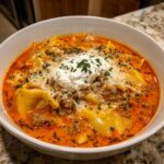 A close-up of a hearty bowl of creamy lasagna soup, featuring pasta, meat sauce, and a dollop of ricotta cheese.