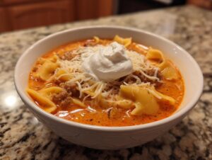 A close-up of a white bowl filled with creamy lasagna soup, featuring pasta, ground meat, shredded cheese, and a dollop of sour cream.