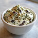 Close-up of a creamy, white olive dip mixed with chopped green herbs and dark olives, served in a small white bowl.