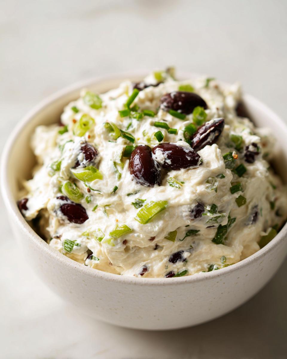Close-up of a creamy white olive dip mixed with chopped black olives and green onions in a speckled white bowl.