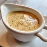 Close-up of creamy, rich peppercorn sauce speckled with black pepper served in a small white gravy boat.