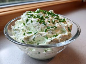 A clear glass bowl filled with creamy pickle dip, garnished generously with chopped fresh chives.