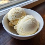 Two scoops of creamy, pale yellow protein ice cream served in a white bowl on a dark wooden surface.
