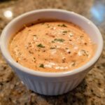 A close-up shot of thick, creamy orange remoulade sauce speckled with green herbs and spices in a small white ramekin.