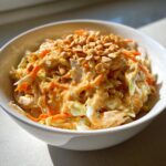 A close-up of a bowl of creamy thai chicken salad mixed with shredded cabbage and carrots, topped generously with chopped peanuts.