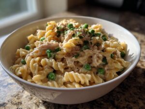 A close-up of creamy tuna pasta mixed with bright green peas, served in a white bowl.