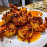 Close-up of perfectly golden-brown, crispy coconut shrimp served on a white plate with a side of bright orange dipping sauce.