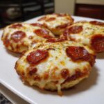 Four freshly baked english muffin pizza halves topped with melted cheese, pepperoni, and herbs on a white plate.