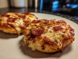 Two perfectly melted pepperoni english muffin pizza halves sitting on a white plate, featuring bubbly cheese and herbs.