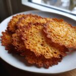 Close-up of several golden brown, delicate oatmeal lace cookies with intricate lacy textures stacked on a light plate.