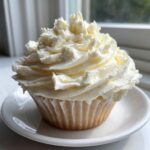 A single vanilla cupcake topped with a generous swirl of fluffy vanilla buttercream frosting, sitting on a small white saucer.