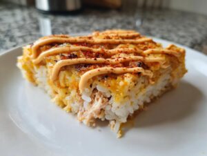 A close-up, featured image of a square slice of baked sushi with layers of rice, creamy topping, and spicy mayo drizzle.