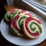 Close-up of four festive red, green, and white pinwheel cookies, coated in sparkling sugar, on a white plate.