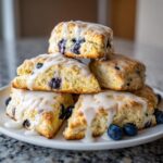 A stack of perfectly flaky blueberry scones drizzled with white glaze, garnished with fresh blueberries.