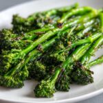 Close-up of bright green, perfectly cooked broccolini stalks glistening with sauce on a white plate.