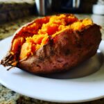 A close-up of a perfectly cooked baked sweet potatoes, split open to show the fluffy, bright orange interior.