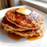 A close-up of a tall stack of golden brown pancakes topped with melting butter and drizzled with syrup, emphasizing fluffy pancakes.