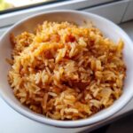 Close-up of fluffy, seasoned Spanish rice served in a white bowl, showing orange-red seasoning and small pieces of onion.