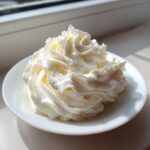 A close-up of a perfect swirl of fluffy vanilla buttercream icing on a small white plate, highlighted by sunlight.