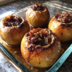 Four glistening baked apples stuffed with a caramelized raisin filling, sitting in syrup in a glass baking dish.