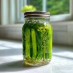 A glass jar filled with fresh cucumbers, dill sprigs, and garlic ready to become refrigerator pickles.
