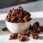 A white bowl overflowing with dark, rich gingerbread granola, featuring visible pecans and dried cranberries.