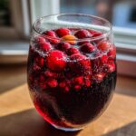 A glass filled with dark red holiday sangria, topped with floating fresh cranberries and a slice of citrus.