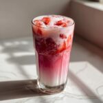 A tall glass filled with layered homemade strawberry milk, featuring crushed strawberries and pink foam.