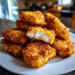 A close-up of crispy, golden homemade chicken nuggets piled on a white plate, one nugget is broken open showing the white chicken inside.
