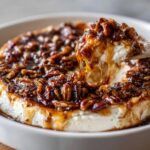 A spoonful of creamy cheese is lifted from a baked pecan pie dip, showing gooey caramel sauce and pecans dripping.