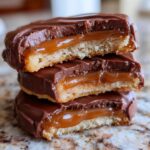A stack of three homemade Twix cookies cut in half, showing the shortbread base, gooey caramel layer, and chocolate coating.