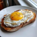 Close-up of delicious egg toast featuring a sunny-side-up egg over creamy ricotta on toasted bread, seasoned with pepper.