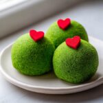 Three bright green, sugar-coated Grinch Oreo Balls, each topped with a small red heart candy, sitting on a white plate.