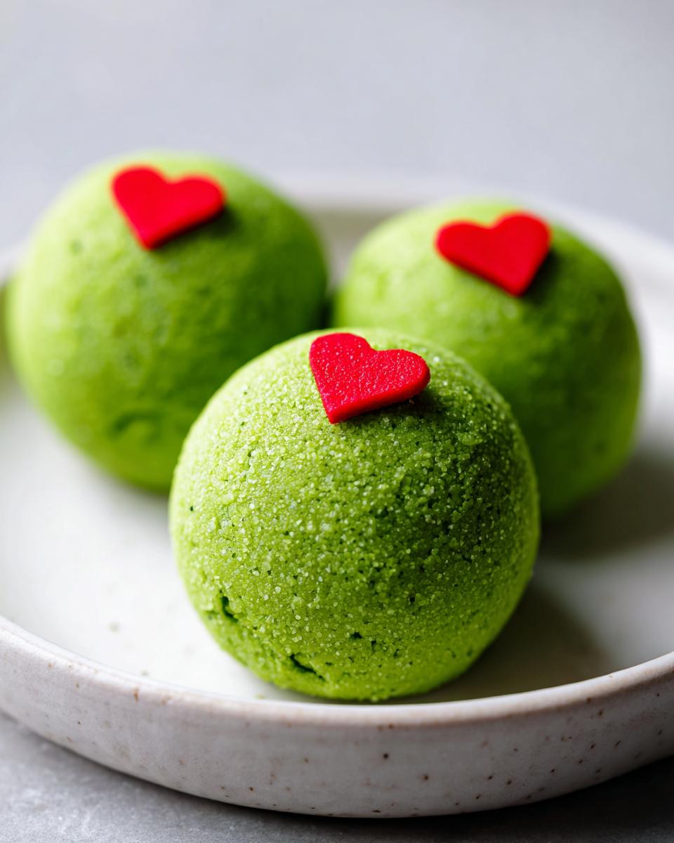 Three bright green Grinch Oreo balls, each topped with a small red heart candy, resting on a white speckled plate.