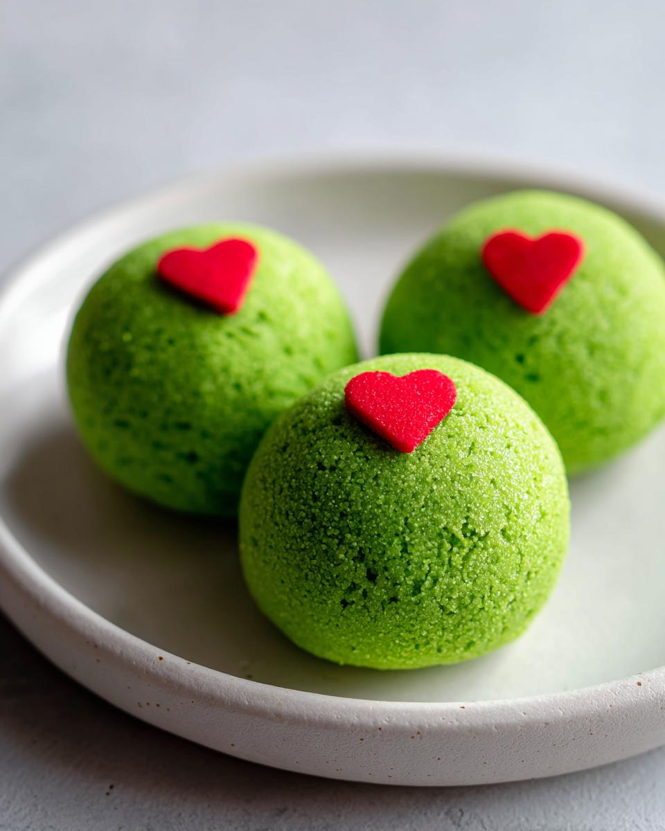 Three bright green Grinch Oreo Balls, dusted with sugar, each topped with a small red heart candy, sitting on a white plate.